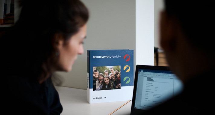 Zwei Schüler sitzen vor einem Laptop und lesen etwas. Im Hintergrund ist ein Ordner mit der Aufschrift Berufswahl-Portfolio zu sehen.