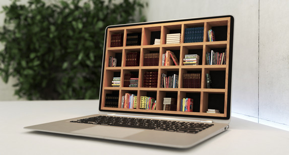 Laptop, der statt einem Monitor ein kleines Bücherregal mit verschiedenen Materialien, Büchern etc. hat, steht auf einem Tisch