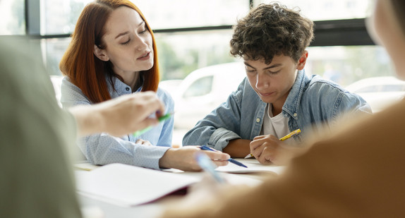 Lehrerin ist in die Hocke gegangen, um einen Shüler im Teeanageralter etwas zu erklären. Mit einem Stift zeigt sie auf eine Stelle in seinem Arbeitsbuch.
