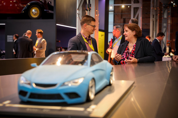 Sportministerkonferenz Sportministerkonferenz 8. und 9. Mai 2025 in Chemnitz Im Vordergrund sieht man ein Miniaturauto, dahinter unterhält sich eine Frau mit einem Mann.