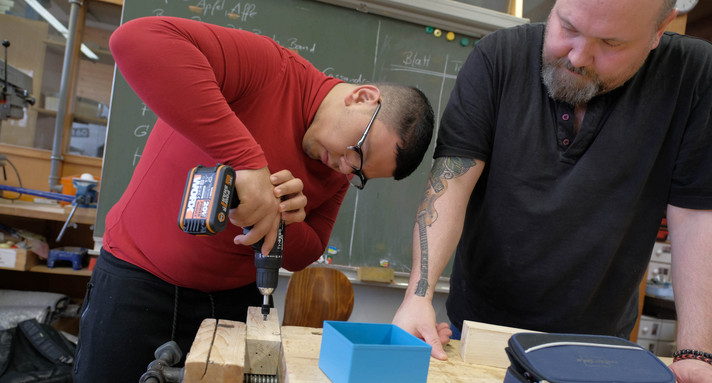 Ein Schüler bohrt mit einem Bohrer ein Loch in ein Stück Holz, ein Lehrer steht daneben