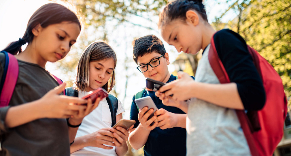 Kinder mit Rucksäcken stehen im Freien und schauen auf ihre Smartphones