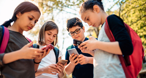 Kinder mit Rucksäcken stehen im Freien und schauen auf ihre Smartphones