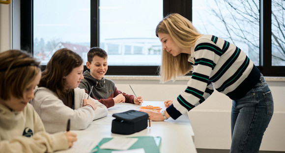 Schülerinnen und Schüler sitzen an Tischen, eine Lehrerin steht ihnen zum Tisch heruntergebeugt gegenüber.