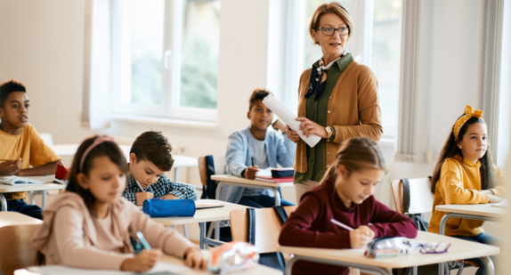 Lehrerin, stehend in der Schulklasse, umgeben von sitzenden Schülerinnen und Schülern