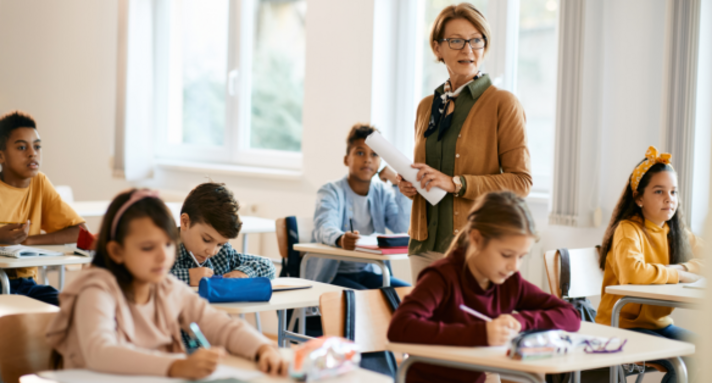 Lehrerin, stehend in der Schulklasse, umgeben von sitzenden Schülerinnen und Schülern