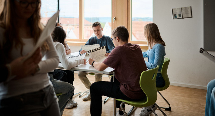 Schülerinnen und Schüler sitzen an einem Tisch und halten Arbeitsblätter in den Händen