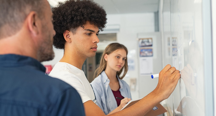 Schüler und Schülerin stehen mit ihrem Lehrer an einem Whiteboard. Der Schüler im Focus schreibt etwas auf das Whiteboard.