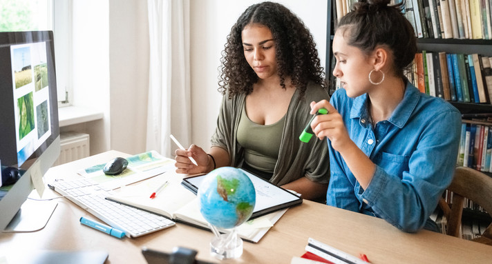 Zwei junge Frauen sitzen mit Stift an einem Schreibtisch und arbeiten an etwas, vor ihnen ist ein Bildschirm und ein Globus zu sehen
