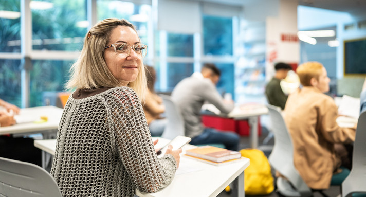 Ältere Schülerin mit lockigen hellen Haaren und Brille schaut im Klassenraum lächelnd über die Schulter in die Kamera.