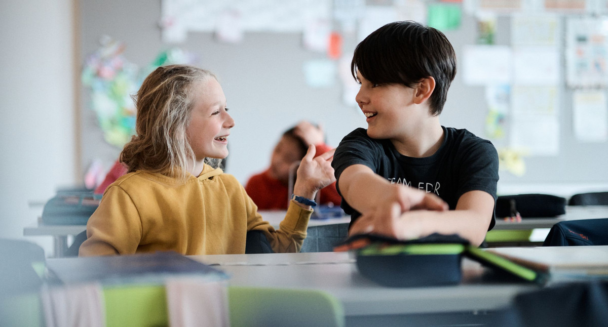 Ein Mädchen und ein Junge sitzen an einem Tisch in der Schule. Sie unterhalten sich miteinander und lachen fröhlich.