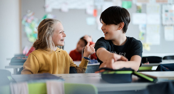 Ein Mädchen und ein Junge sitzen an einem Tisch in der Schule. Sie unterhalten sich miteinander und lachen fröhlich.