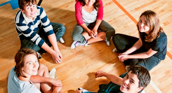 Vier Jugendliche - drei Mädchen und zwei Jungen - sitzen im Kreis in einer Sporthalle.  