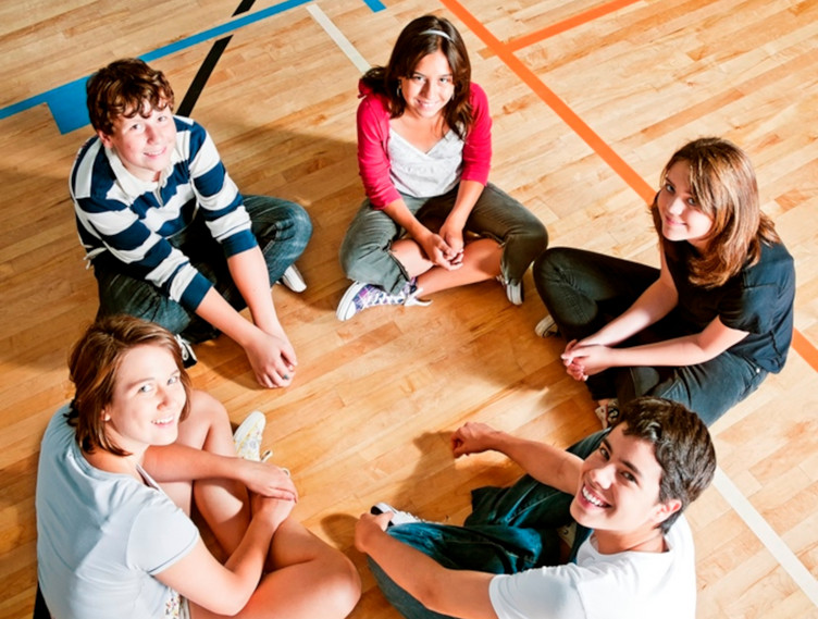 Vier Jugendliche - drei Mädchen und zwei Jungen - sitzen im Kreis in einer Sporthalle.  