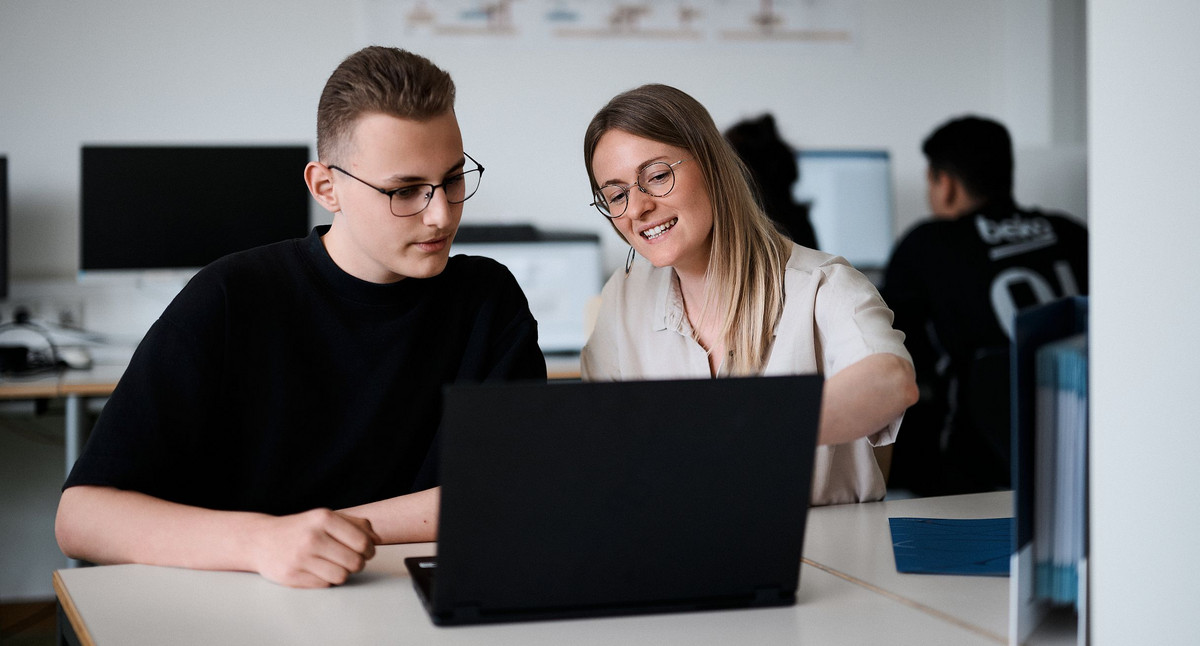 Ein Mann und eine Frau sitzen vor einem Laptop. Die Frau zeigt mit dem Finger auf etwas auf dem Bildschirm.