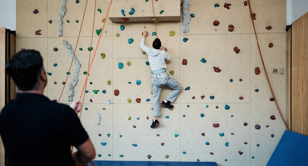 Ein Jugendlicher an einer Kletterwand. Eine weitere Person sichert ihn mit einem Seil.
