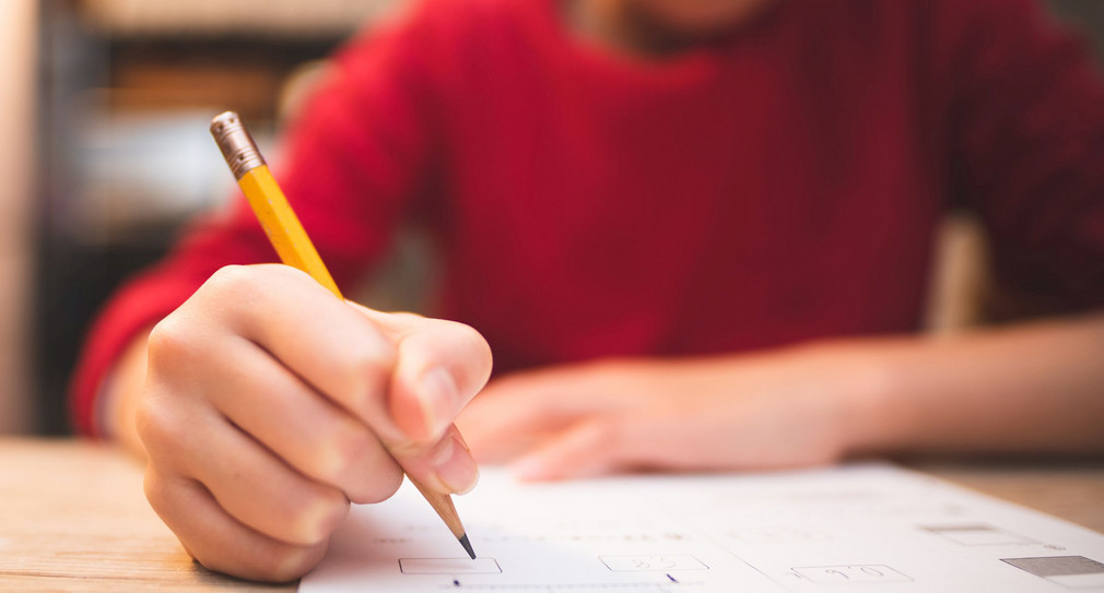 eine jugendliche Person sitzt mit einem Stift an der Hand an einem Tisch und löst eine schriftliche aufgabe auf dem Papier vor ihr.