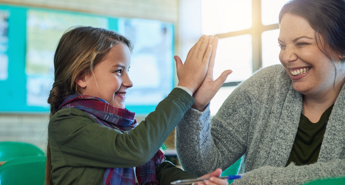 Eine Grundschülerin und eine Lehrerin in einem Schulraum klatschen sich ab