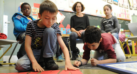 Klassenraum mit einer Lehrerin, drei Schülern und einer Schülerin. Im Hintergrund eine Tafel. Zwei Schüler legen etwas auf dem Boden, die anderen schauen zu.