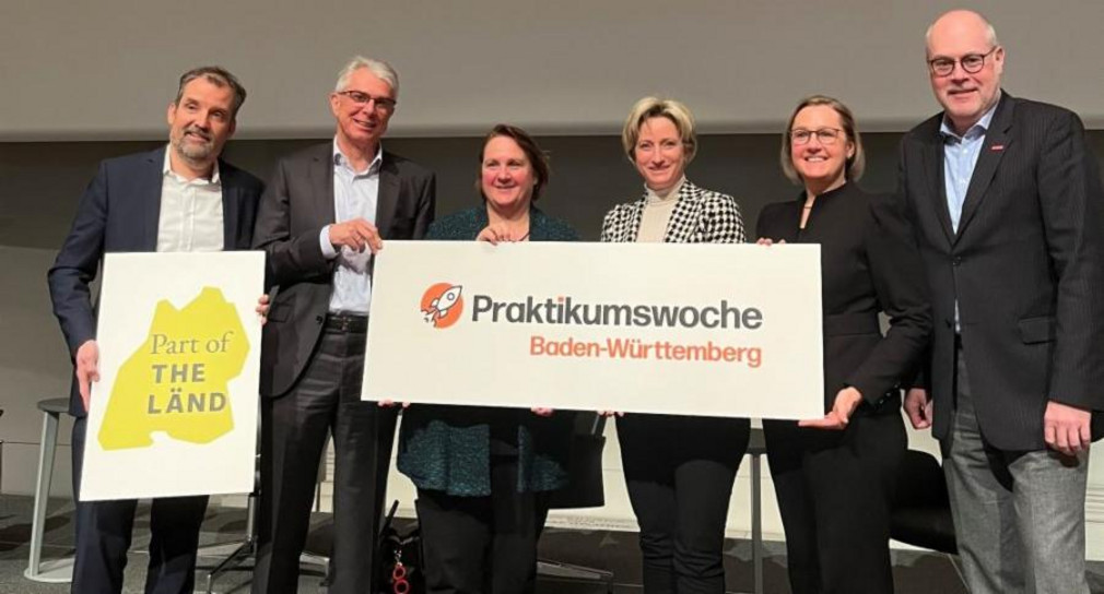 Sechs Personen stehen nebeneinander und halten ein großes Schild hoch. 