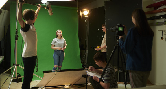 Medienklasse vor grünem Hintergrund beim Film drehen. Ein Schüler hält das Mikrofon hoch. Alles richtet sich auf die jugendliche Moderatorin aus.