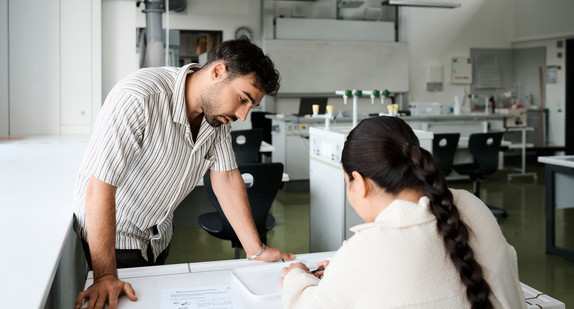 Eine Schülerin sitzt an einem Tisch in einem Chemieraum. Sie arbeitet mit diversen Utensilien. Eine Lehrkraft steht neben dem Tisch und unterstützt sie.
