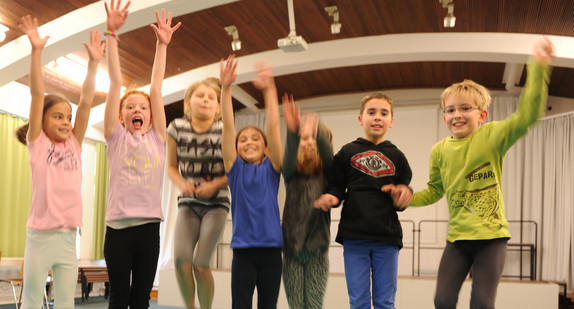 Mehrere Grundschülerinnen und Grundschüler springen lachend in einer Reihe in die Luft und recken die Arme nach oben