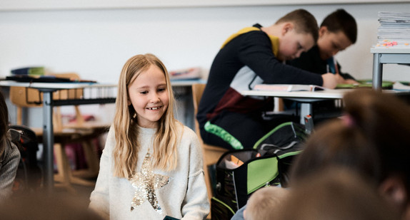 Ein etwa elfjähriges Mädchen sitzt im Sitzkreis auf dem Boden eines Klassenzimmers. Im Hintergrund sitzen zwei Klassenkameraden an einem Tisch und bearbeiten Übungsaufgaben.