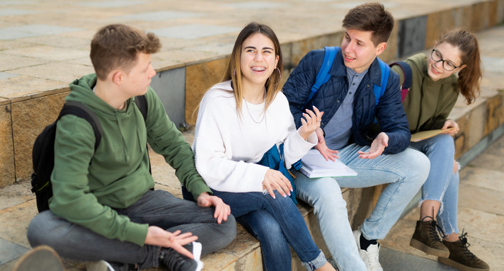 Zwei jugendliche Schülerinnen und zwei Schüler sitzen nebeneinander auf einer hohen und breiten Treppenstufe aus Sandstein und reden miteinander. Eine der Jugendlichen scheint eine Idee zu haben.