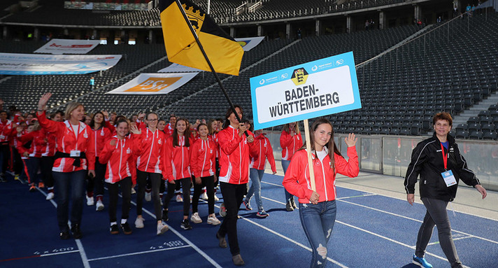 Eine Gruppe von jugendlichen Sportlern geht mit Fahne in einem Sportstadion im Kreis. 