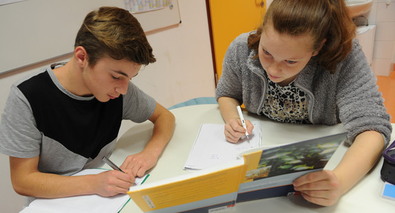 Eine Schülerin und ein Schüler sitzen zusammen an einem Tisch und schreiben in ihr Heft. Das Mädchen hält dabei ein geöffnetes Lehrbuch - für beide sichtbar - in einer Hand 