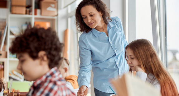 Lehrerin weist auf etwas im Arbeitsbuch einer Schülerin im Teenageralter. An dem Tisch davor sitzt ein Schüler und lernt.