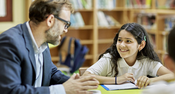 Lehrer erklärt dunkelhaarigem lächelnden Mädchen etwas an einem Tisch sitzend