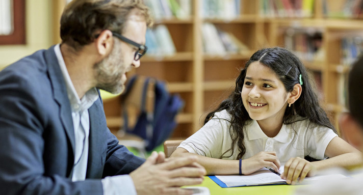 Lehrer erklärt dunkelhaarigem lächelnden Mädchen etwas an einem Tisch sitzend