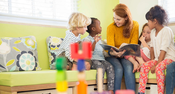 Eine Frau sitzt zwischen vier Kindern auf einer Bank und liest ihnen aus einem Buch vor