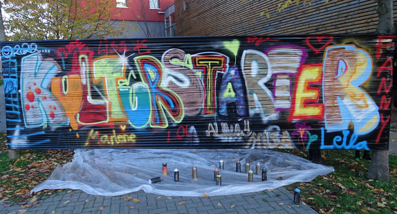 Vor einer Mauer stehen Farbspraydosen auf dem Boden, auf der Mauer steht in bunten Buchstaben "Kulturstarter". 