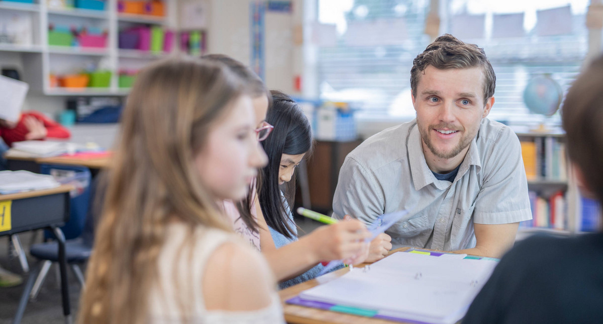 Ein Lehrer sitzt bei Schülerinnen und Schülern an einem Tisch in einem Schulraum