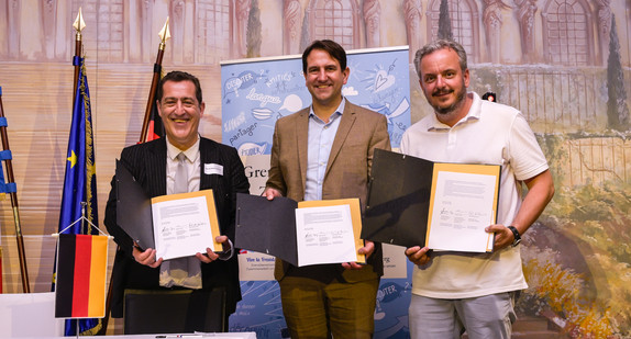 Drei Männer stehen vor einer Wand und halten jeweils ein Blatt Papier in der Hand.