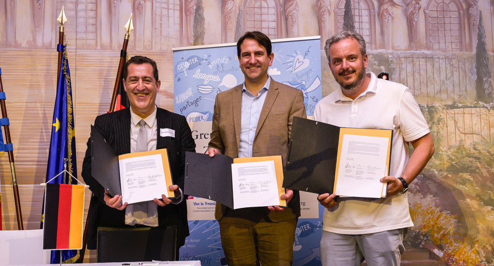 Drei Männer stehen vor einer Wand und halten jeweils ein Blatt Papier in der Hand.
