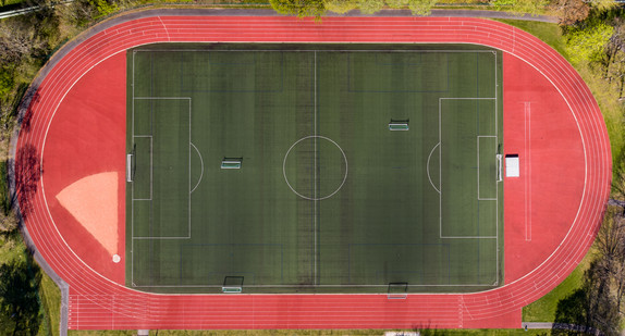 Eine Aufnahme von oben auf einen grünen Fussballplatz, der umrandet ist von einer roten Laufbahn. 