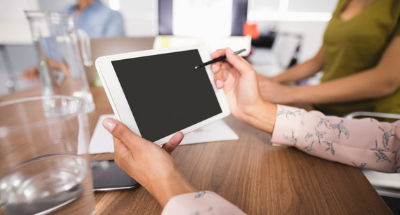 Die Hände einer Frau halten ein Tablet. Im Hintergrund weitere Personen an einem Tisch.