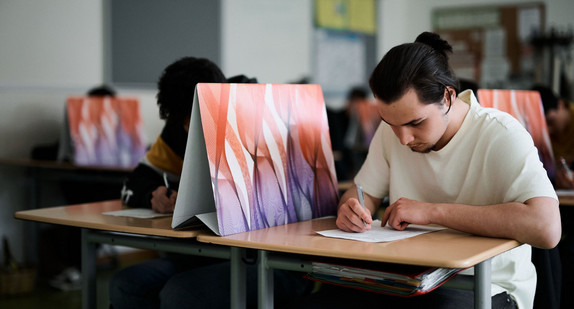 Das Bild zeigt mehrere jugendliche Schülerinnen und Schüler in einem Klassenraum. Sie sitzen an Tischen und bearbeiten Aufgaben. Zwischen Ihnen sind Sichtschutze aus Pappe aufgebaut.