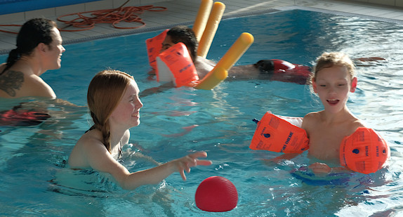 Drei Kinder mit Lehrer im Schwimmbecken. Zwei Kinder tragen Schwimmhilfen an den Armen, eins davon arbeitet mit zwei Poolnudeln