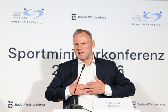 Sportministerkonferenz Sportministerkonferenz 8. und 9. Mai 2025 in Chemnitz Ein Mann mit weißen Hemd und blauem Jacket steht an einem Rednerpult, hinter sich eine weiße Wand mit dem logo der Sportministerkonferenz.