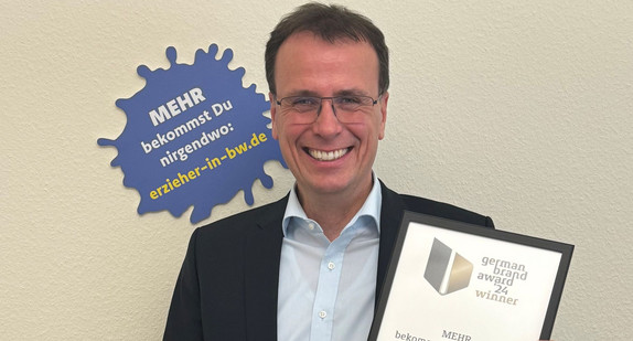 Vor gelbem Hintergrund steht ein Mann undn lächert, er hält eine Urkunde in der Hand.  
