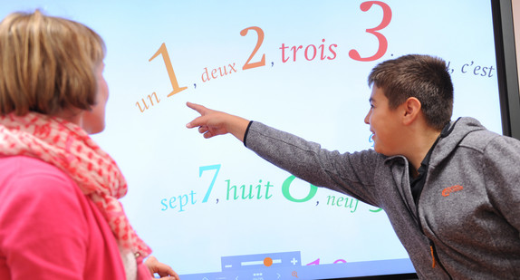 Lehrerin und Schüler stehen vor einem Whiteboard, die Ziffern mit französicher Bezeichnung zeigt. Der Schüler weist auf die 1.