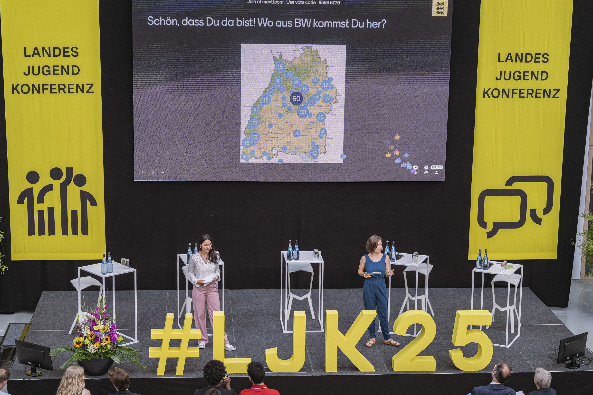 Auf einer Bühne stehen zwei junge Frauen mit Mikrofonen in der Hand. 