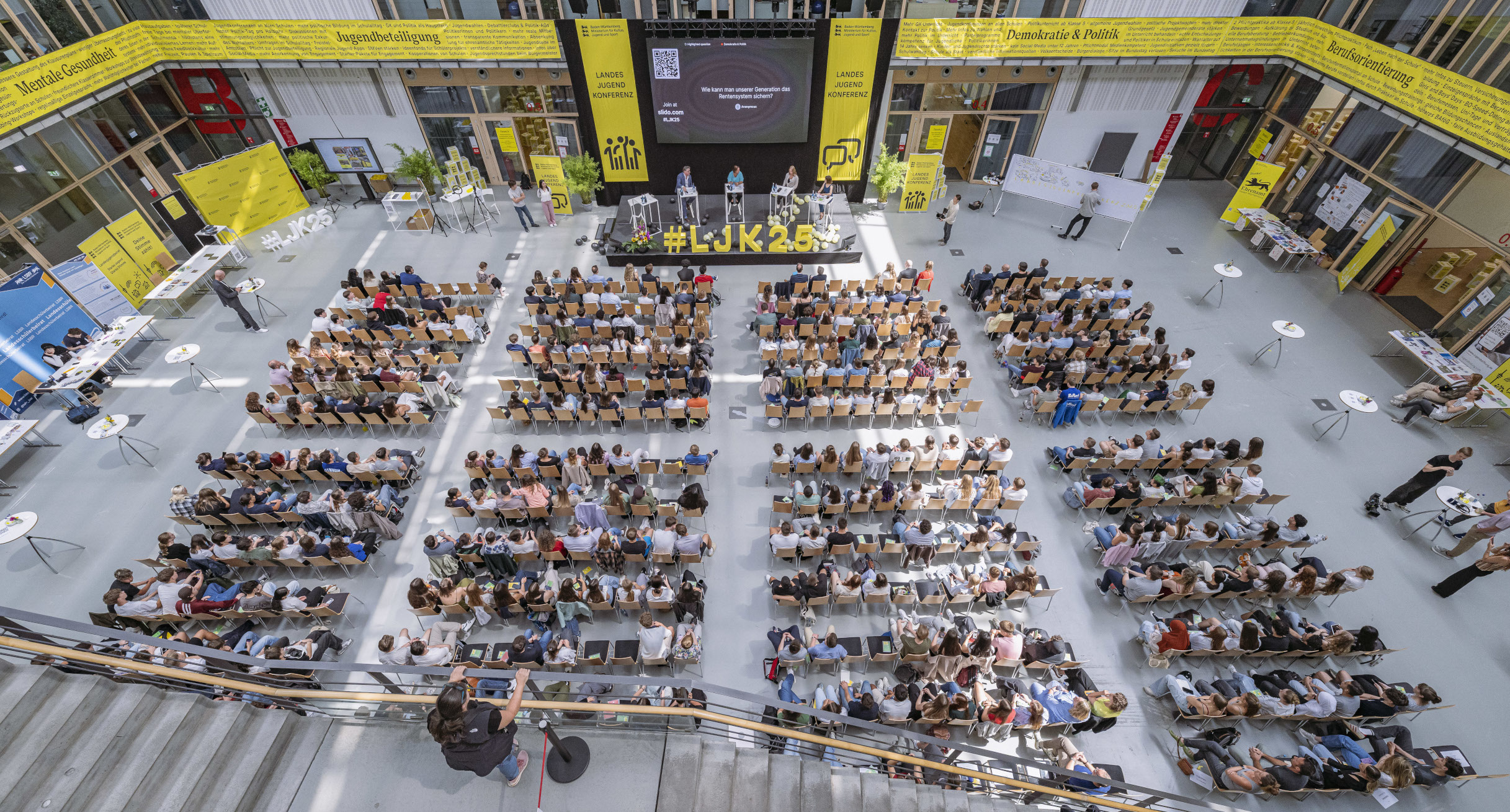 Von oben sieht man in eine Halle herab, in der viele Personene auf Stühlen sitzen. 